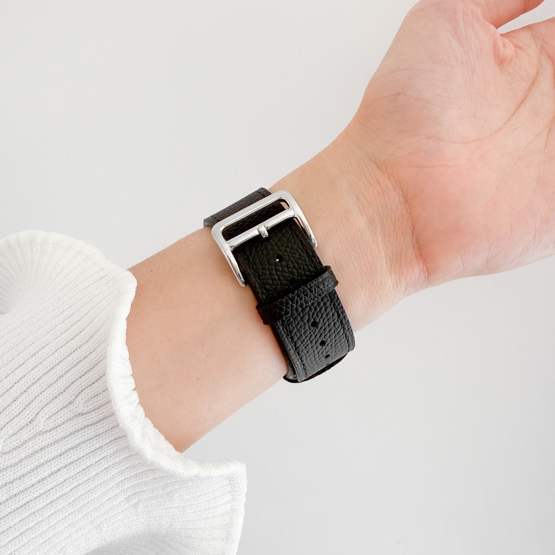 A close-up of a black textured leather watch strap with a silver buckle worn on a person's wrist, with a white ribbed sweater sleeve visible.