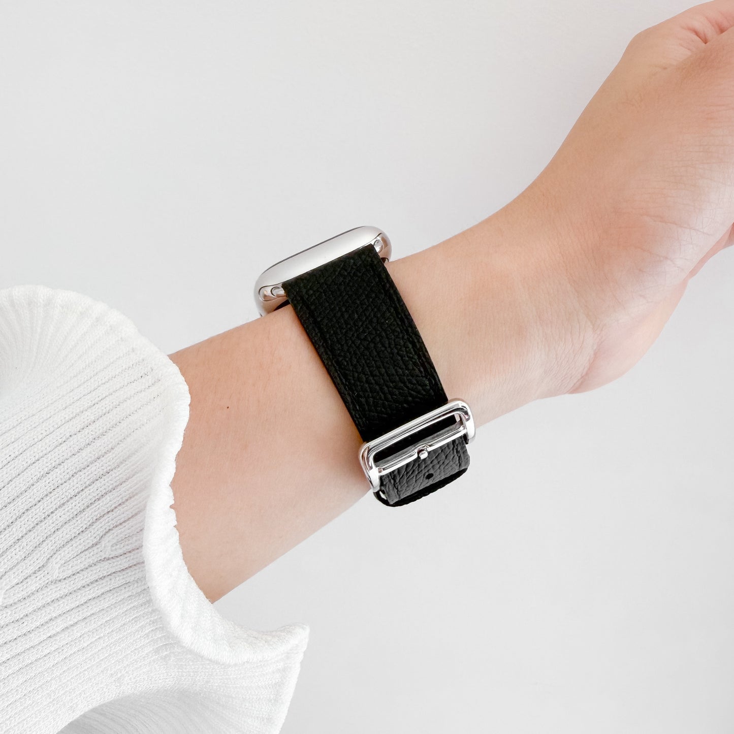A close-up of a person's wrist wearing a black textured leather watch band with silver metal hardware, paired with a white ribbed knit sleeve against a light gray background.
