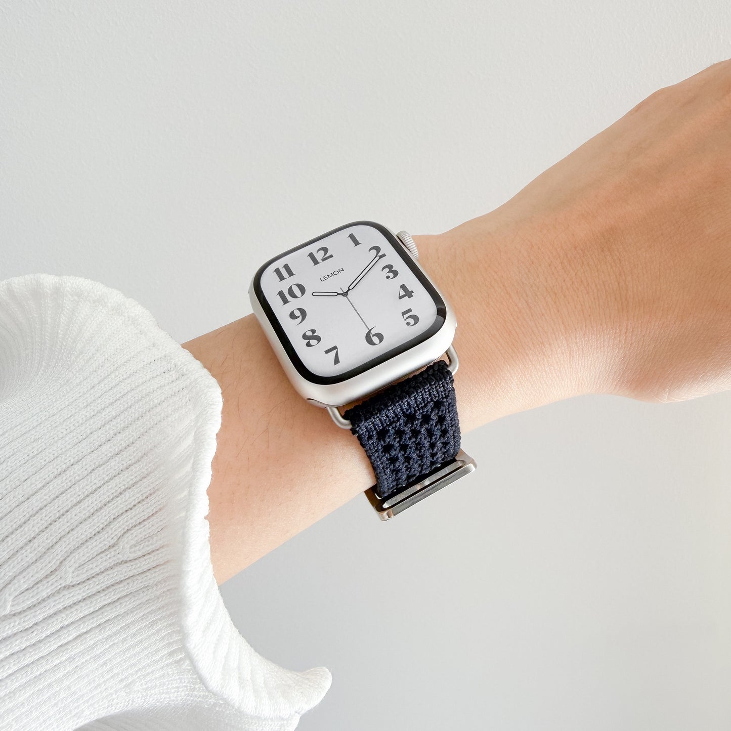 A close-up of a smartwatch with a apple watch and a dark navy woven fabric strap, worn on a person's wrist against a light gray background.
