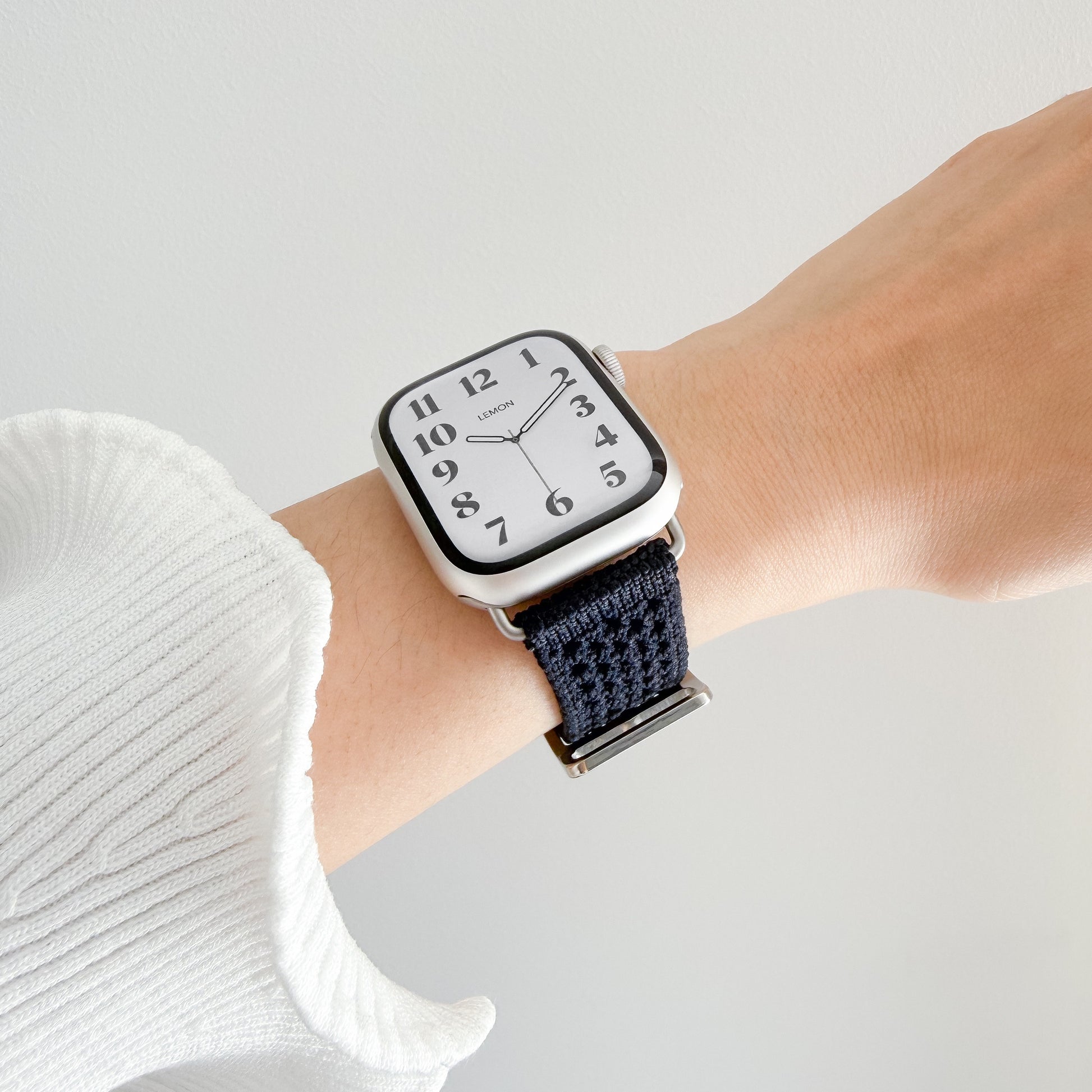 A close-up of a smartwatch with a apple watch and a dark navy woven fabric strap, worn on a person's wrist against a light gray background.