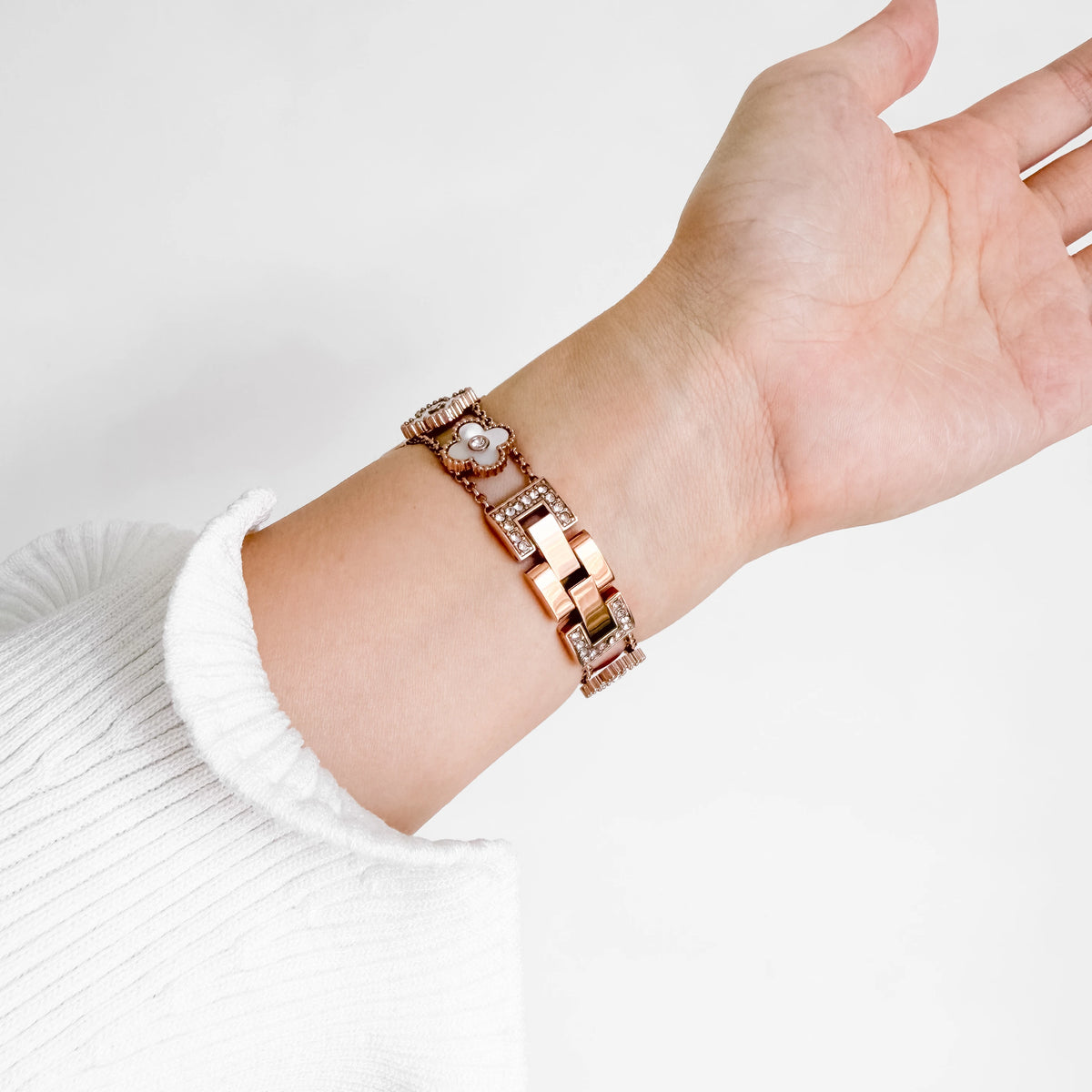 A close-up of a person's wrist wearing a rose gold link bracelet with a central pearl and crystal details, paired with a white ribbed knit sleeve.