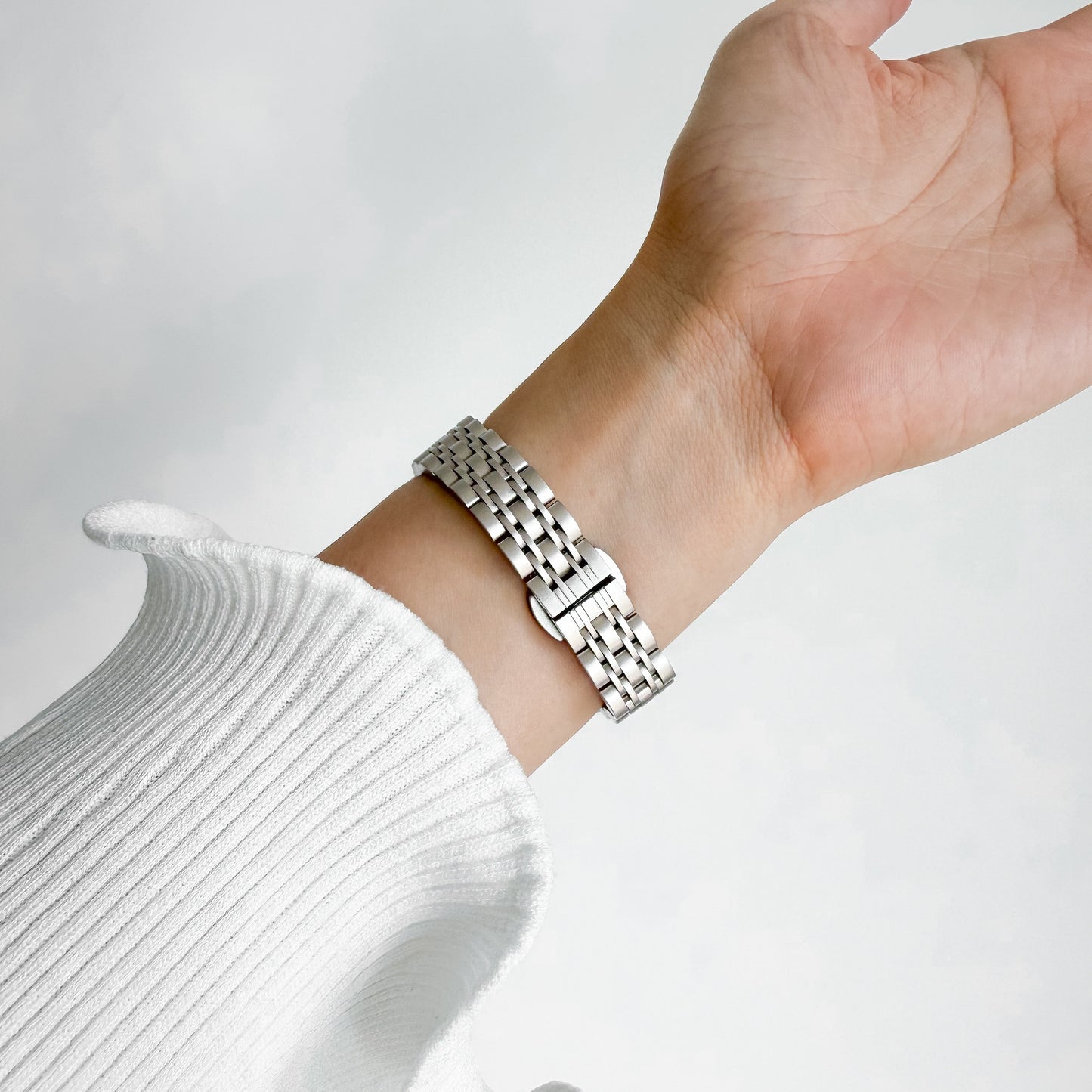 A close-up of a starlight stainless steel link watch band worn on a person's wrist, paired with a white ribbed knit sweater sleeve, set against a soft, light gray background.