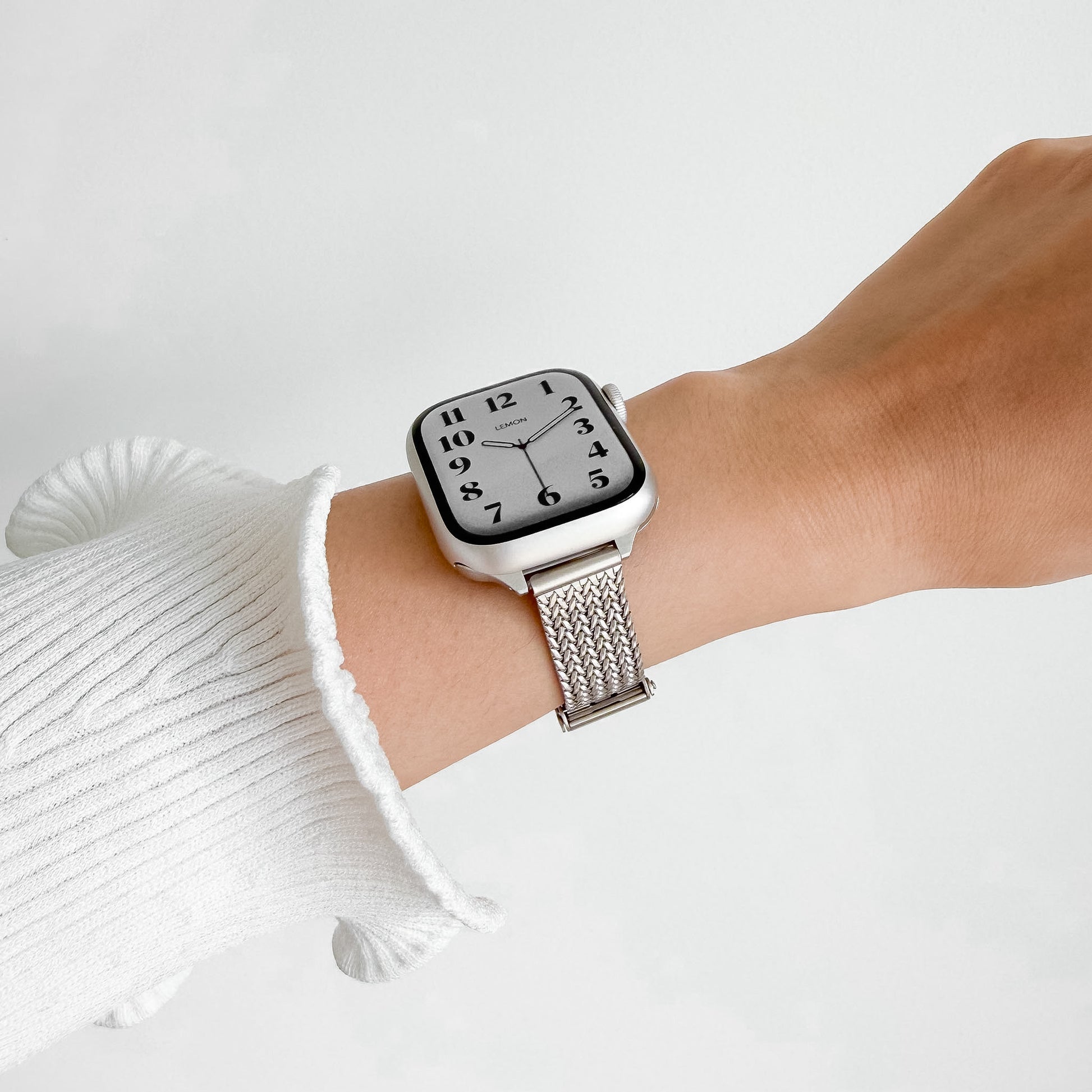 A silver smartwatch with a square black-and-white analog-style face and a silver mesh band, worn on a person's wrist. The person is wearing a white ribbed knit sleeve against a clean light gray background.